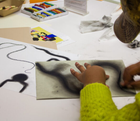 High Museum of Art offers free admission for all A woman sits at a table surrounded by oil pastels. She wears a hat and green sweater and is drawing large, swirling black lines with an oil pastel on paper.