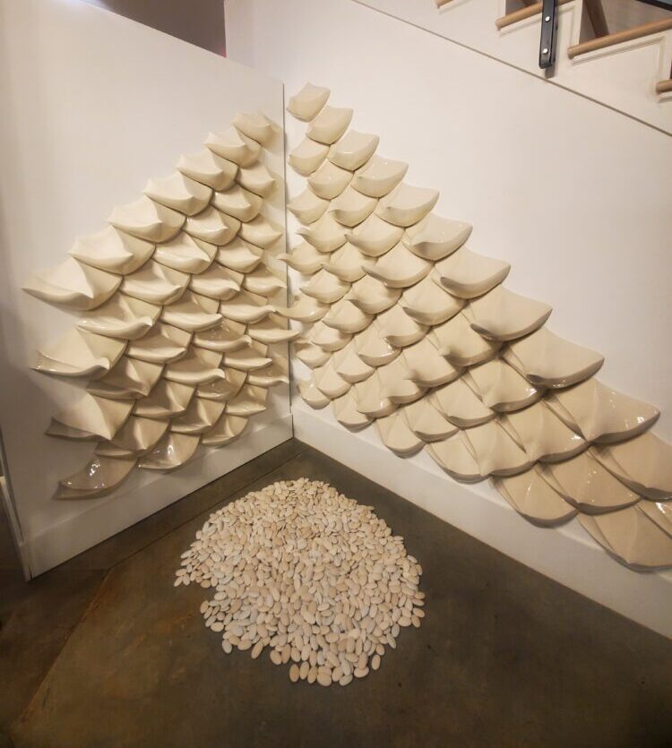 Cream-colored sculptures shaped like pinecone scales cover two walls that form a corner. The floor in the corner has a circular pile of pebbles.