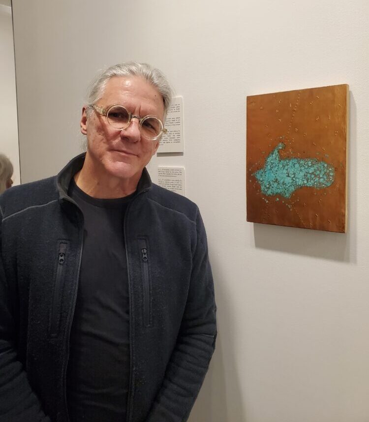 A white man with gray hair, glasses, a black shirt, and a black jacket stands to the left of an art piece. The piece is small and rectangular. It is brown with a turquoise shape in the center. The piece has braille detailing.