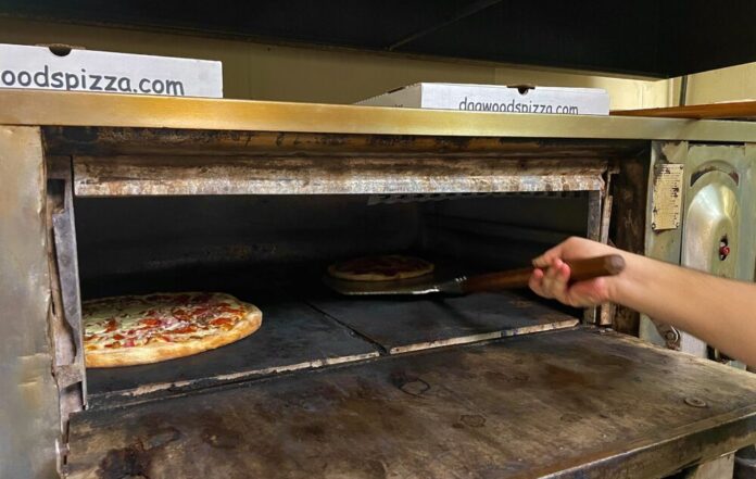 A hand holding a pizza peel scoops a pizza out of a large oven. A second pepperoni pizza bakes in the oven to the left of the first one.