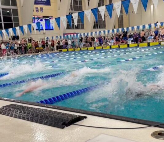 Georgia high school swimmers make waves at state championship Wide shot of swimmers racing in a large pool. The lanes are divided by blue and white barriers. The swimmers are all underwater, splashing and approaching the finish. Blue and white triangular flags hang above the pool. A crowd cheers from the sidelines.