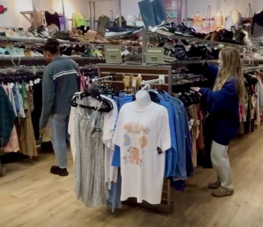Why more shoppers are opting for thrift store savings A girl with long blonde hair wearing a navy blue sweater sorts through a rack of shirts at a thrift store. To her left, a mannequin torso displays a graphic tee. On top of the clothing rack are rows of sneakers and boots for sale.
