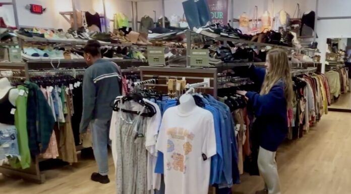 Why more shoppers are opting for thrift store savings A girl with long blonde hair wearing a navy blue sweater sorts through a rack of shirts at a thrift store. To her left, a mannequin torso displays a graphic tee. On top of the clothing rack are rows of sneakers and boots for sale.