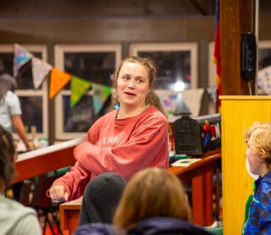 From camper to staff: How one camp’s mission changes lives A woman with tied-back brown hair wearing a red sweatshirt dances with a group of children. The kids are dressed in casual clothing.
