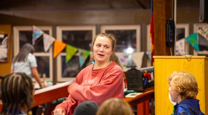 From camper to staff: How one camp’s mission changes lives A woman with tied-back brown hair wearing a red sweatshirt dances with a group of children. The kids are dressed in casual clothing.