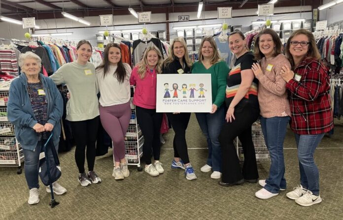 Foster care foundation: Nine women stand posing in a line smiling at the camera. Behind them are racks of hanging clothing. The two women in the middle are holding a sign featuring a graphic of children that reads 
