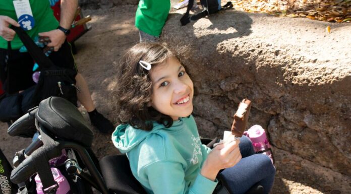 Adventure of a lifetime for Kennesaw girl with chronic illness A young girl with short hair wearing an aqua hoodie sits in a wheelchair and smiles up at the camera. She is holding a Mickey Mouse ice cream bar.