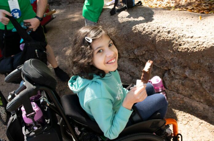 A young girl with short hair wearing an aqua hoodie sits in a wheelchair and smiles up at the camera. She is holding a Mickey Mouse ice cream bar.