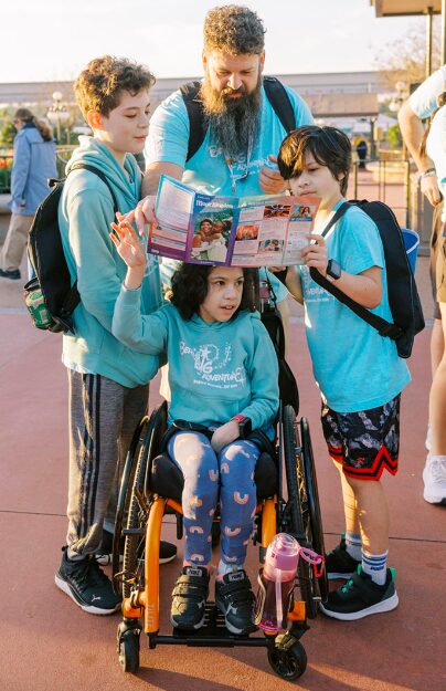 Four people stand as a group looking at a paper map. In the middle stands a man with a long beard holding the map. To his left and right are two young boys. In the center is a young girl in a wheelchair. The four are all wearing matching Bert's Big Adventure shirts.