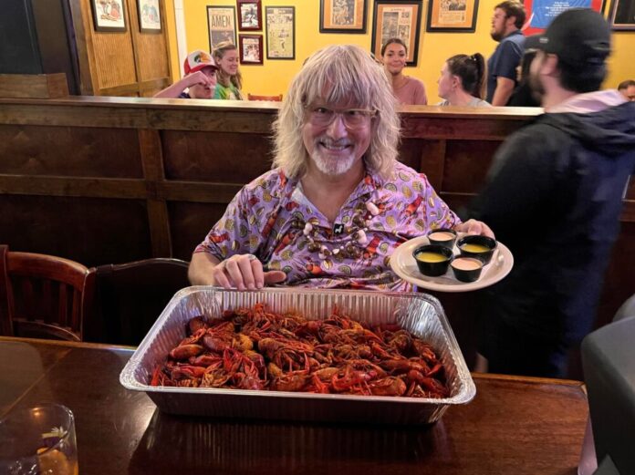 An older man with long, wavy, gray hair and bangs smiles at the camera. He has glasses and a gray goatee. He is holding a tray of dipping sauces in his left hand and sitting at a table with a seafood boil in an aluminum tray.