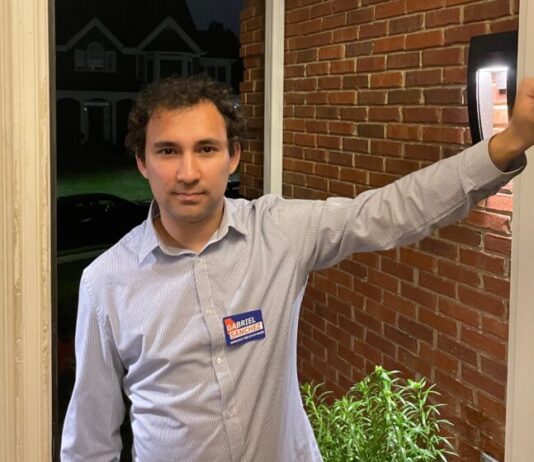 Georgia House candidate offers inside look at campaign process A man knocking on an open door with a blue shirt. He is smiling and is wearing a campaign tag. The background is a brick house and a dark sky.