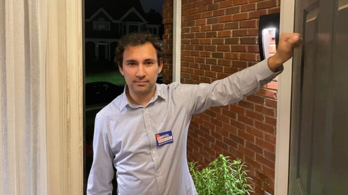 A man knocking on an open door with a blue shirt. He is smiling and is wearing a campaign tag. The background is a brick house and a dark sky.