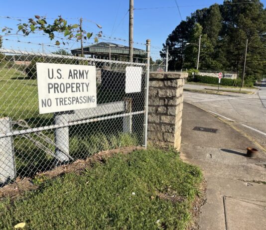 Gov. Kemp limits land purchases near military bases A white sign with black text reads "U.S. Army Property No Trespassing." It is posted on a barbed wire fence. The background is street corner and blue sky.