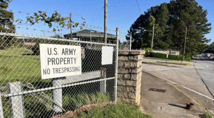 Gov. Kemp limits land purchases near military bases A white sign with black text reads "U.S. Army Property No Trespassing." It is posted on a barbed wire fence. The background is street corner and blue sky.