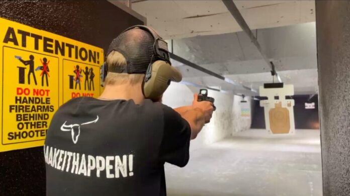 A man shoots a pistol in a gun range. He is facing away from the camera and wearing a baseball hat, black t-shirt, and large headphones. He is aiming the pistol at a target. A large yellow sign reads 