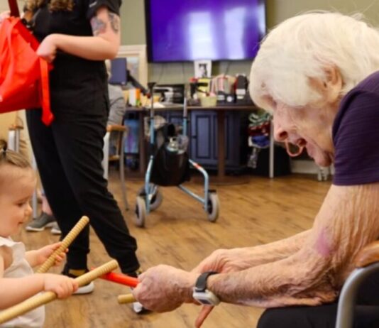 Unique music therapy program bridges generations On the left, a toddler-aged girl wearing a white top with light hair in an updo holds a set of rhythm sticks. On the right, an elderly woman with short white hair and a purple top sits in a chair with another pair of rhythm sticks. The two are smiling at each other.