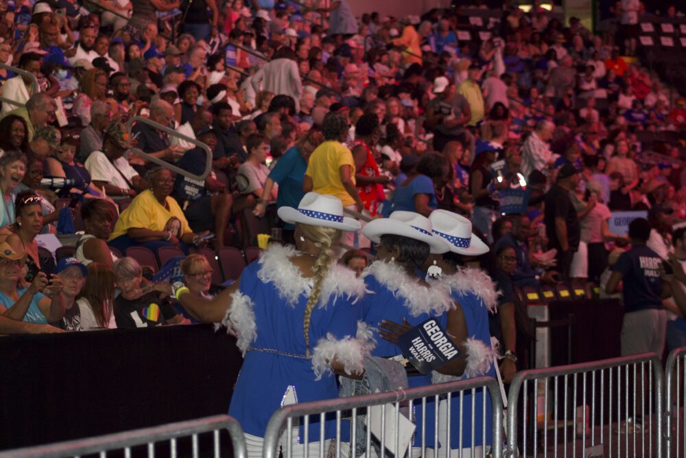 Three women wearing cowboy hats and blue tops trimmed with white feathers stand in a row pressed against a metal baricade. Their backs are to the camera and they're facing a large crowd in a stadium. The crowd is washed out in red lighting.