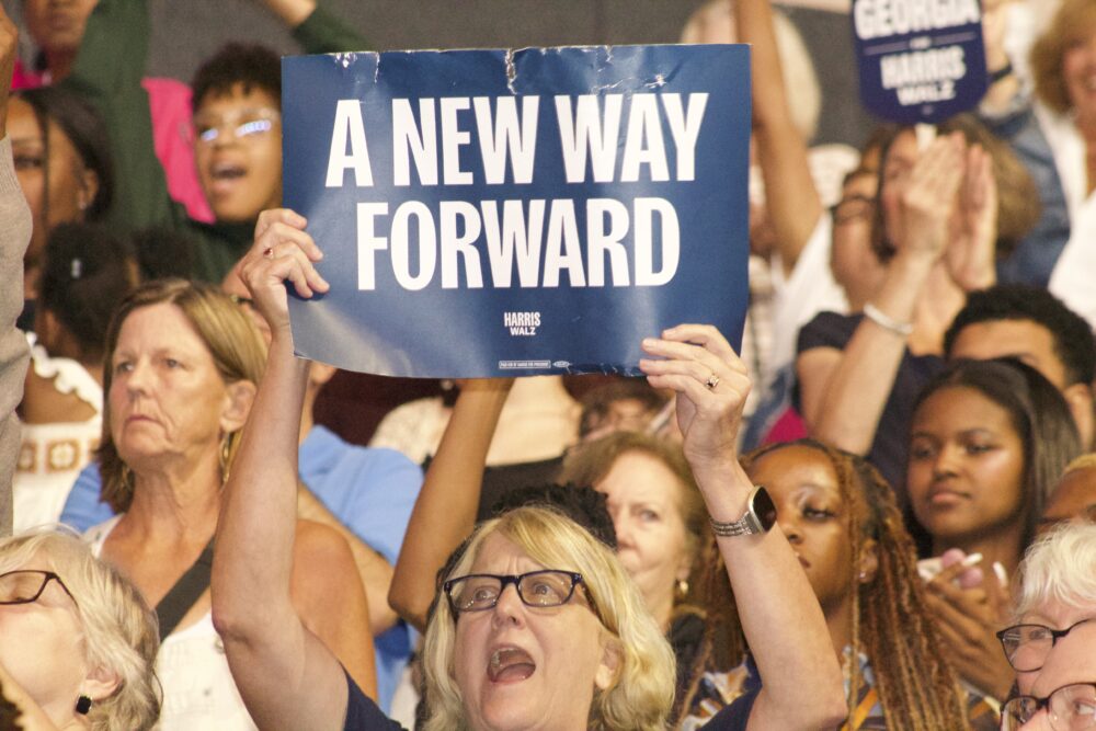 A woman stands in a crowd, yelling and holding a blue sign that reads, "A New Way Forward."