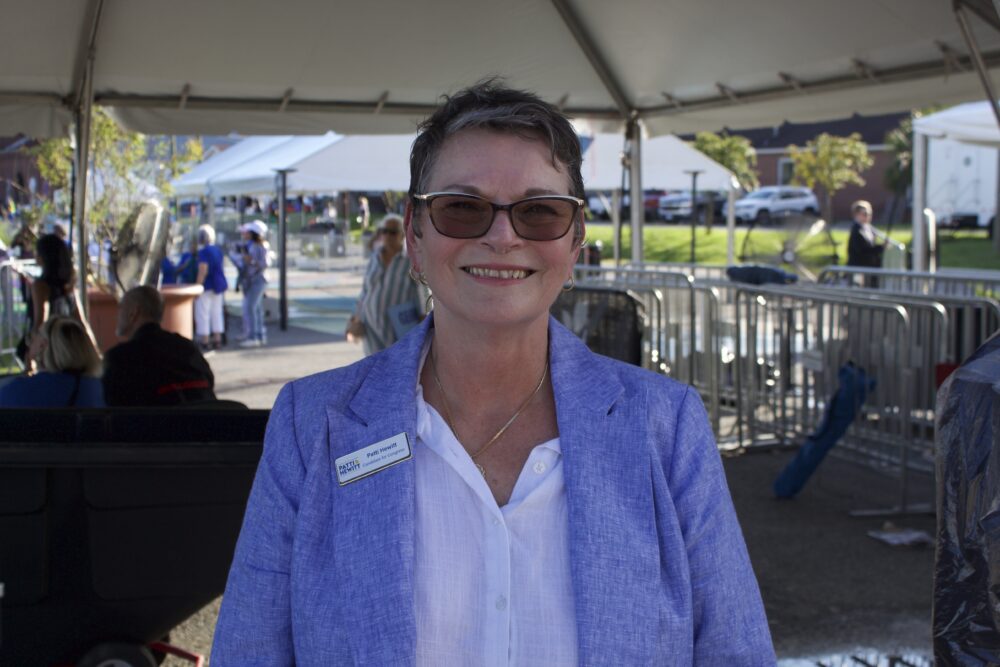 A white woman with short, spiky gray hair wearing sunglasses and a gray blazer smiles at the camera. She is standing in the center of the frame and is the only subject. 