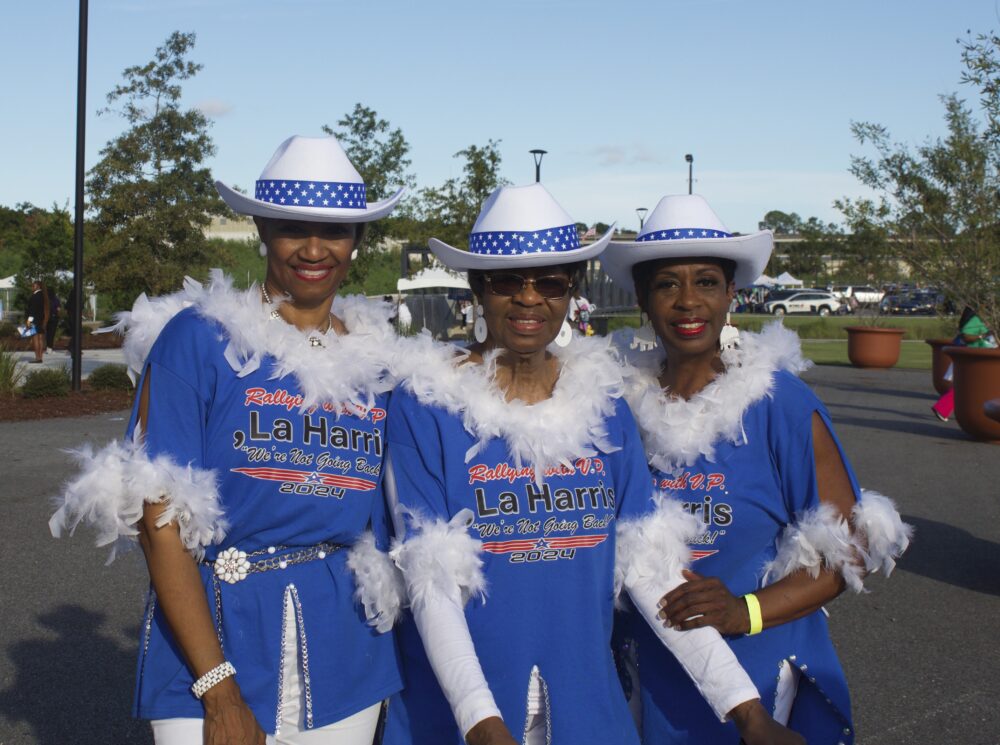 Three Black women stand in a row, posing and smiling at the camera. They are all wearing white cowboy hats and blue tops trimmed with white feathers. The shirts read, "Rallying with V.P. La Harris, "We're Not Going Back," 2024." The woman in the middle is wearing a white long-sleeved shirt underneath, as well as sunglasses. All three women are wearing large, white elephant earrings.