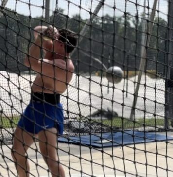 Kennesaw hammer thrower returns home after challenging Olympic Games A shirtless white man wearing blue shorts and sunglasses is standing behind a net, swinging a metal ball attached to a wire, known as a hammer.