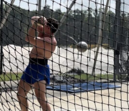 Kennesaw hammer thrower returns home after challenging Olympic Games A shirtless white man wearing blue shorts and sunglasses is standing behind a net, swinging a metal ball attached to a wire, known as a hammer.