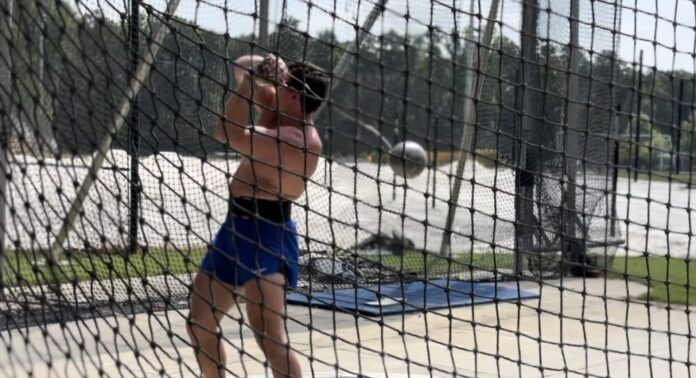 A shirtless white man wearing blue shorts and sunglasses is standing behind a net, swinging a metal ball attached to a wire, known as a hammer.