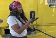 Are truckers finally on the “Highway to Wellness?” A Black woman with long black hair, a gray t-shirt, and a pink silk hair scarf stands next to a large yellow truck, filling up the truck tank with gasoline. She is looking off to the right and gesturing with her right hand, which is gloved.