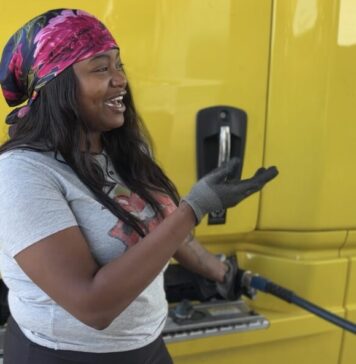 Are truckers finally on the “Highway to Wellness?” A Black woman with long black hair, a gray t-shirt, and a pink silk hair scarf stands next to a large yellow truck, filling up the truck tank with gasoline. She is looking off to the right and gesturing with her right hand, which is gloved.