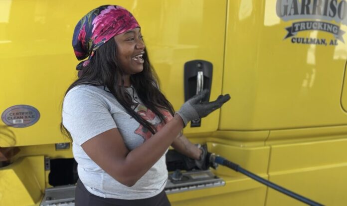 A Black woman with long black hair, a gray t-shirt, and a pink silk hair scarf stands next to a large yellow truck, filling up the truck tank with gasoline. She is looking off to the right and gesturing with her right hand, which is gloved.