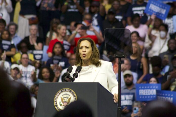 Vice President Kamala Harris stands speaking at a podium. She is wearing a white top and blazer. A large crowd of people is gathered behind her, out of focus in the camera.