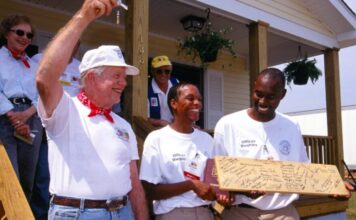 Jimmy Carter, former president and humanitarian, dies at age 100 Former president Jimmy Carter stands on the porch of a home holding a key in the air. Next to him, two Black men stand smiling, holding a plank of wood covered in signatures. Behind Carter stand his wife, Rosalynn.