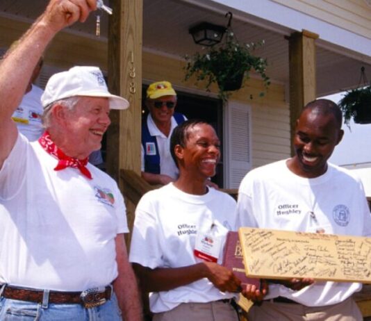 Jimmy Carter, former president and humanitarian, dies at age 100 Former president Jimmy Carter stands on the porch of a home holding a key in the air. Next to him, two Black men stand smiling, holding a plank of wood covered in signatures. Behind Carter stand his wife, Rosalynn.