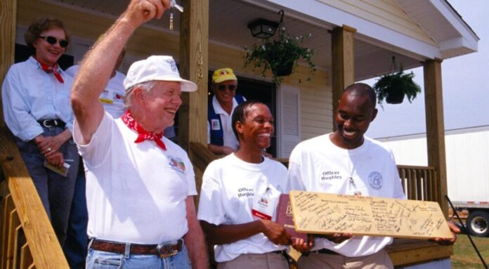 Former president Jimmy Carter stands on the porch of a home holding a key in the air. Next to him, two Black men stand smiling, holding a plank of wood covered in signatures. Behind Carter stand his wife, Rosalynn.