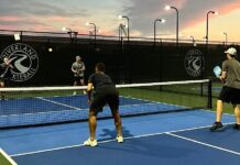Kennesaw State’s pickleball team is a smashing success Four men stand on a pickleball court, two standing on each side of the low net. The men in focus are facing away from the camera, holding rackets and wearing dark workout clothing. The sunset is purple and pink in the distance.