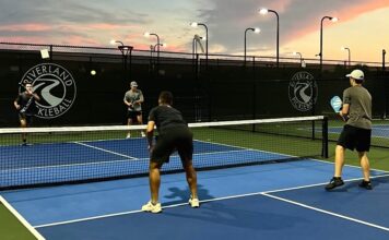 Kennesaw State’s pickleball team is a smashing success  Four men stand on a pickleball court, two standing on each side of the low net. The men in focus are facing away from the camera, holding rackets and wearing dark workout clothing. The sunset is purple and pink in the distance.