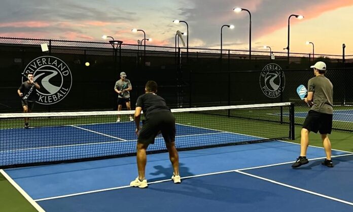 Four men stand on a pickleball court, two standing on each side of the low net. The men in focus are facing away from the camera, holding rackets and wearing dark workout clothing. The sunset is purple and pink in the distance.