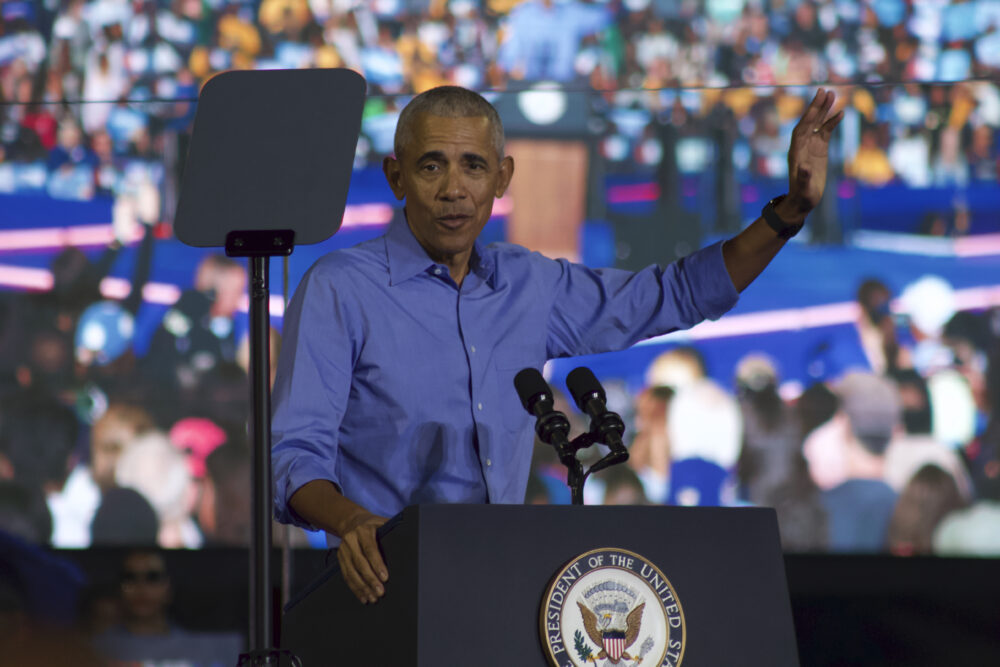 Barack Obama wears a blue button-up shirt and stands at a podium. He is speaking and gesturing with his left hand. 