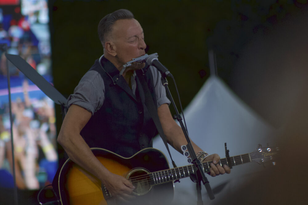 Bruce Springsteen wears a grey shirt and a dark vest. He is singing, blowing into a harmonica and playing guitar. 