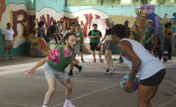 A man with a pink shirt and a green penny jersey stands with bent knees in front of a Black nonbinary person holding a basketball. A crowd of people watches the game about to start.