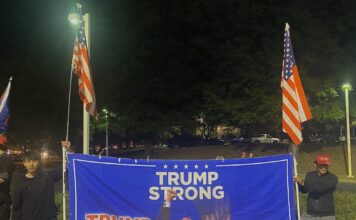 Trump holds rally in Duluth to encourage voting Two Japanese men wearing all black hold American flags and a large blue sign with a photo of Donald Trump after he was shot. The text reads, "Trump strong, Chosen by God."