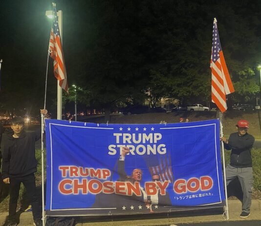 Trump holds rally in Duluth to encourage voting Two Japanese men wearing all black hold American flags and a large blue sign with a photo of Donald Trump after he was shot. The text reads, "Trump strong, Chosen by God."
