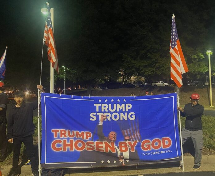 Two Japanese men wearing all black hold American flags and a large blue sign with a photo of Donald Trump after he was shot. The text reads, 