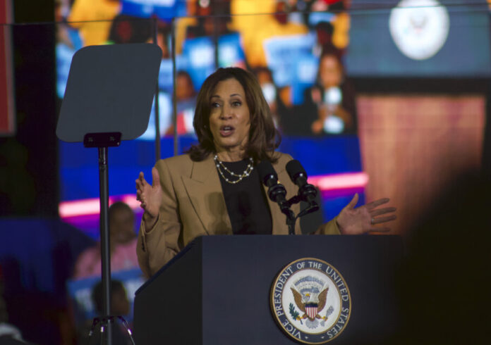 Kamala Harris stands behind a podium giving a speech. She is wearing a black top and tan blazer.