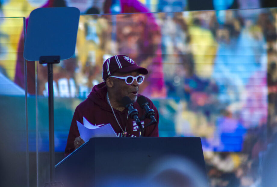 Spike Lee stands at a podium giving a speech to a large crowd. He is wearing a maroon hat and sweatshirt, both reading "Morehouse College," and he is wearing white sunglasses.