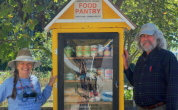 Canton charity stocks pantries for families in need Two older people stand to the sides of a yellow wooden food pantry. To the left of the pantry is a white woman with gray hair in a braid, a blue shirt, and a visor. To the right of the pantry is a taller white man with a baseball hat, long gray hair and a gray beard, and a blue button up shirt.