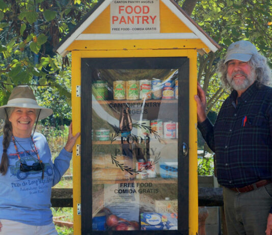 Canton charity stocks pantries for families in need Two older people stand to the sides of a yellow wooden food pantry. To the left of the pantry is a white woman with gray hair in a braid, a blue shirt, and a visor. To the right of the pantry is a taller white man with a baseball hat, long gray hair and a gray beard, and a blue button up shirt.