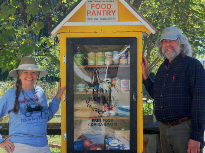 Two older people stand to the sides of a yellow wooden food pantry. To the left of the pantry is a white woman with gray hair in a braid, a blue shirt, and a visor. To the right of the pantry is a taller white man with a baseball hat, long gray hair and a gray beard, and a blue button up shirt.