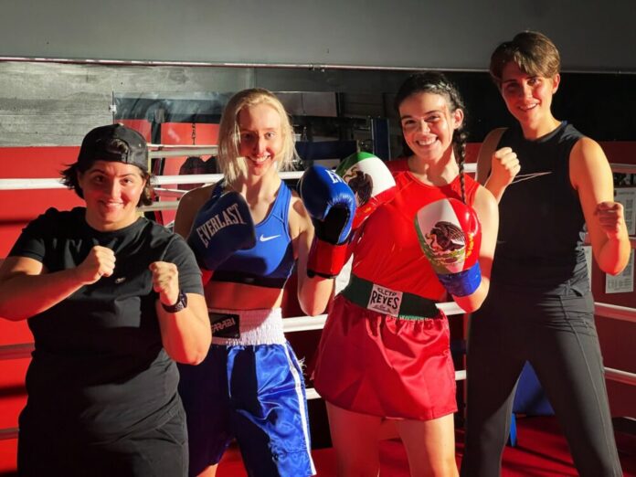 Four women stand with their fists raised, smiling at the camera. On the far left, the shortest woman wears a backwards hat and a black t-shirt. To her right is a blonde woman wearing a matching blue workout set and blue boxing gloves. Next to her is a girl with blood on her temple wearing a red matching set with braids and boxing gloves of the Mexican flag. Lastly, on the far right, a tall woman with short hair wears a black tank top and leggings.