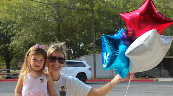 The faces of Fulton County voters on Election Day A blonde white woman wearing a white t-shirt and sunglasses kneels on the ground. Her right arm is wrapped around her daughter, a young girl with long hair and a pink dress, and her left hand is holding three balloons, red, white, and blue stars. Both people are wearing Georgia "I Voted" stickers.